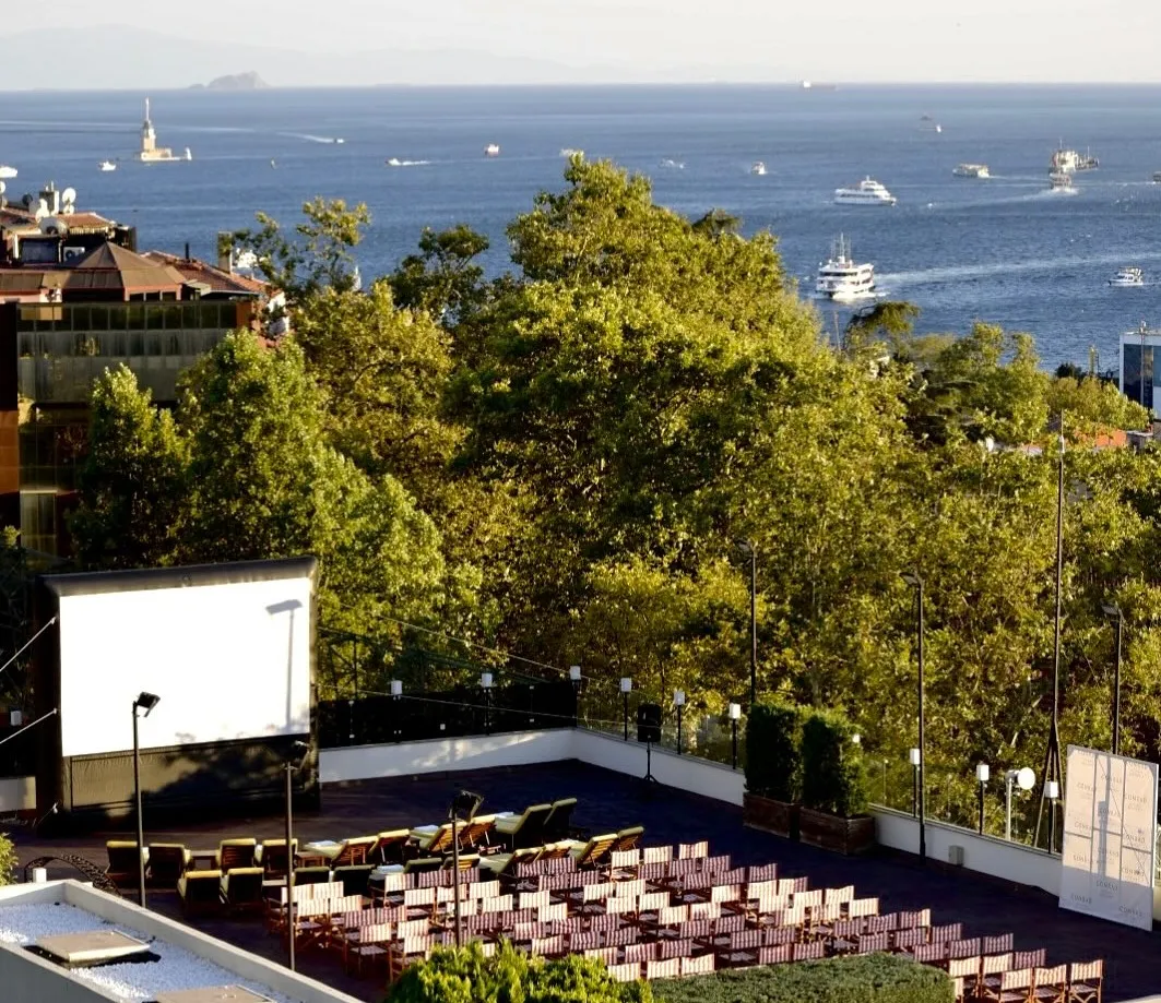Conrad İstanbul Bosphorus'un Aura Garden'ındaki açık hava sinemasında, Boğaz'ın ve uzakta Kız Kulesi'nin görüldüğü yemyeşil ağaçlarla çevrili terasta beyaz bir perde önünde film izlemek için düzenlenmiş çok sayıda sandalye ve şezlong yer alıyor.