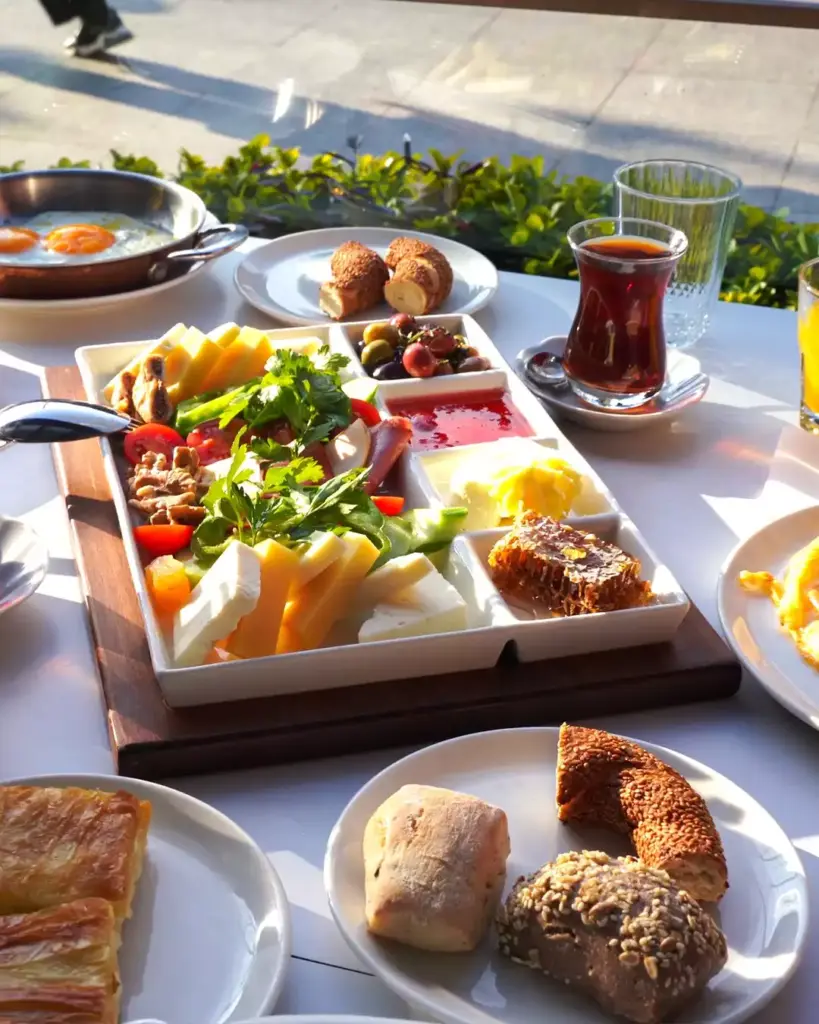 Güneşli bir ortamda beyaz bir masa üzerinde özenle hazırlanmış, peynir çeşitleri, zeytin, taze yeşillikler, reçel, bal peteği, sahanda yumurta, simit, börek ve çay gibi zengin içeriğe sahip, göz alıcı bir Türk kahvaltısı sergileniyor.