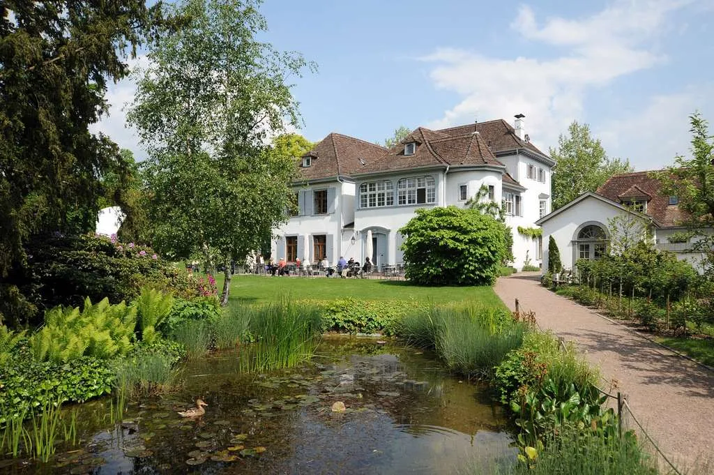 Park Riehen'deki Restaurant Beyeler'de huzurlu bir dış mekan sahnesi; kahverengi çatılı ve yeşil panjurlu, beyaz, geleneksel tarzda bir bina, yemyeşil çimler, ağaçlar ve nilüfer yaprakları ile bir ördeğin bulunduğu bir göletle çevrili, hepsi açık mavi bir gökyüzünün altında.