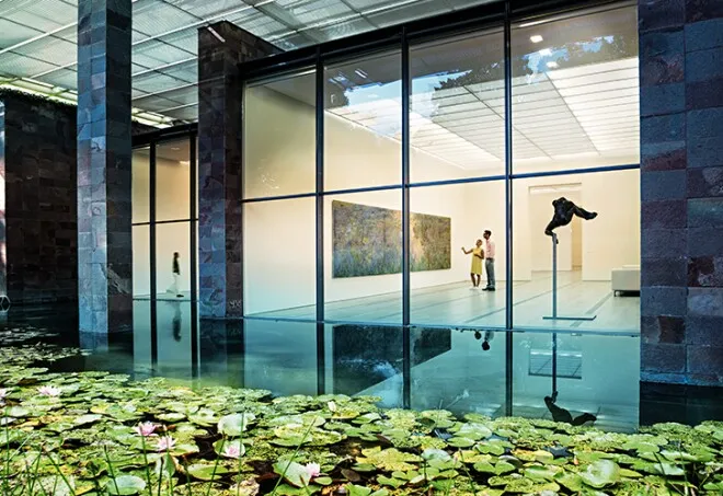 Fondation Beyeler müzesinin dışından, büyük cam pencerelerden aydınlık bir sergi alanına bakış. İki ziyaretçi soyut bir tabloyu ve koyu renkli, heykelsi bir sanat eserini inceliyor. Ön planda, nilüferli yansıtıcı bir gölet huzurlu atmosfere katkıda bulunuyor.