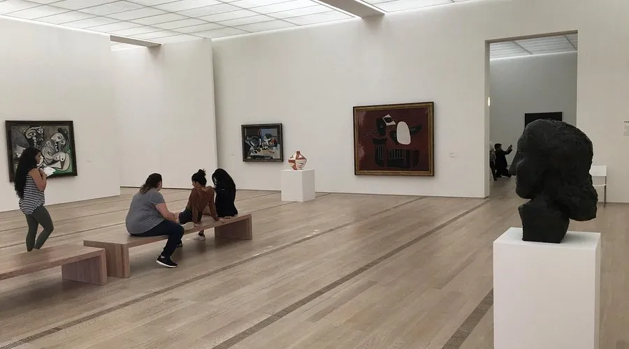 Fondation Beyeler'in içinde geniş, iyi aydınlatılmış bir sanat galerisi; açık renk ahşap zeminler ve beyaz duvarlar. Solda bir Picasso tablosu da dahil olmak üzere birkaç tablo sergileniyor ve ön planda beyaz bir kaide üzerinde koyu renk bir büst heykeli bulunuyor. Ziyaretçiler mekanın çeşitli yerlerine dağılmış durumda, bazıları bir bankta oturuyor.
