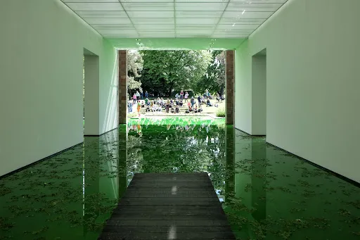 Fondation Beyeler'deki sürükleyici bir sanat enstalasyonu, muhtemelen Olafur Eliasson'a ait; tavanı yansıtan büyük, yeşilimsi tonlu bir su havuzu içeren bir iç mekanı sergiliyor ve uzakta insanların toplandığı parlak yeşil bir çayıra açılıyor. Ahşap bir iskele ön plandan suya doğru uzanıyor.