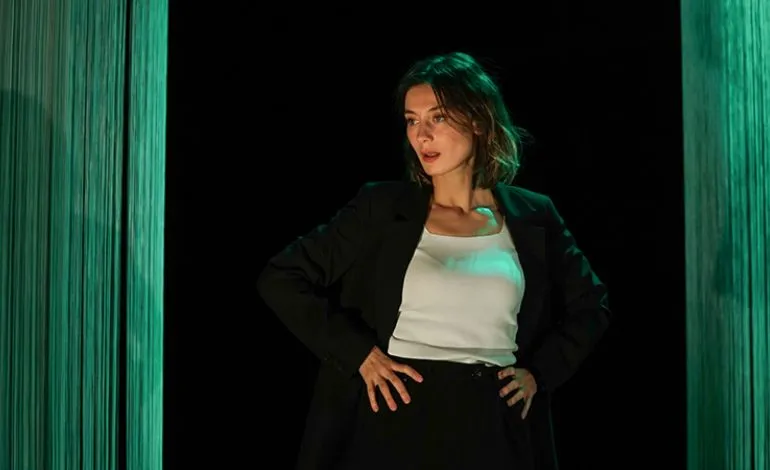 Sahne ışıklarıyla aydınlanmış, üzerinde siyah ceket ve beyaz bir bluz bulunan, elleri belinde hafifçe sola dönük poz veren bir kadın oyuncu, 27. Yapı Kredi Afife Tiyatro Ödülleri için aday gösterilen tiyatro oyunlarından birinden etkileyici bir sahneyi sergiliyor.