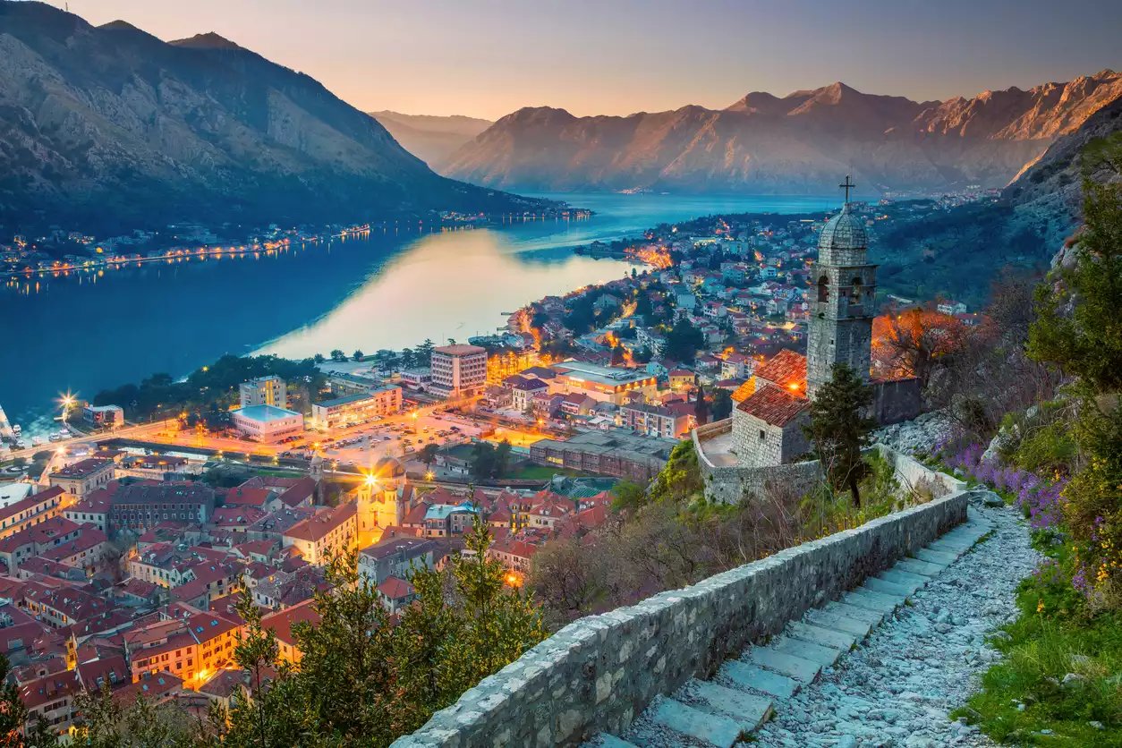 Kotor Körfezi'nin masmavi sularına nazır, dağların çevrelediği tarihi Karadağ şehrinin ışıklarla aydınlanan binalarını, yamaca kıvrılarak yükselen taş patika ve tepedeki kilise kulesini gün batımının pastel renklerinde gösteren büyüleyici bir manzara.