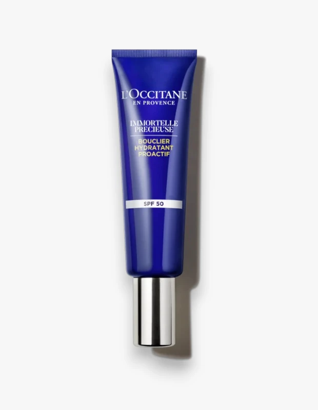 Beyaz bir arka plan üzerinde dikey duran, alt kısmında gümüş renkli bir kapak ve üzerinde SPF 50 yazısı bulunan koyu lacivert L'Occitane Immortelle Précieuse nemlendirici UV kalkanı tüpü.