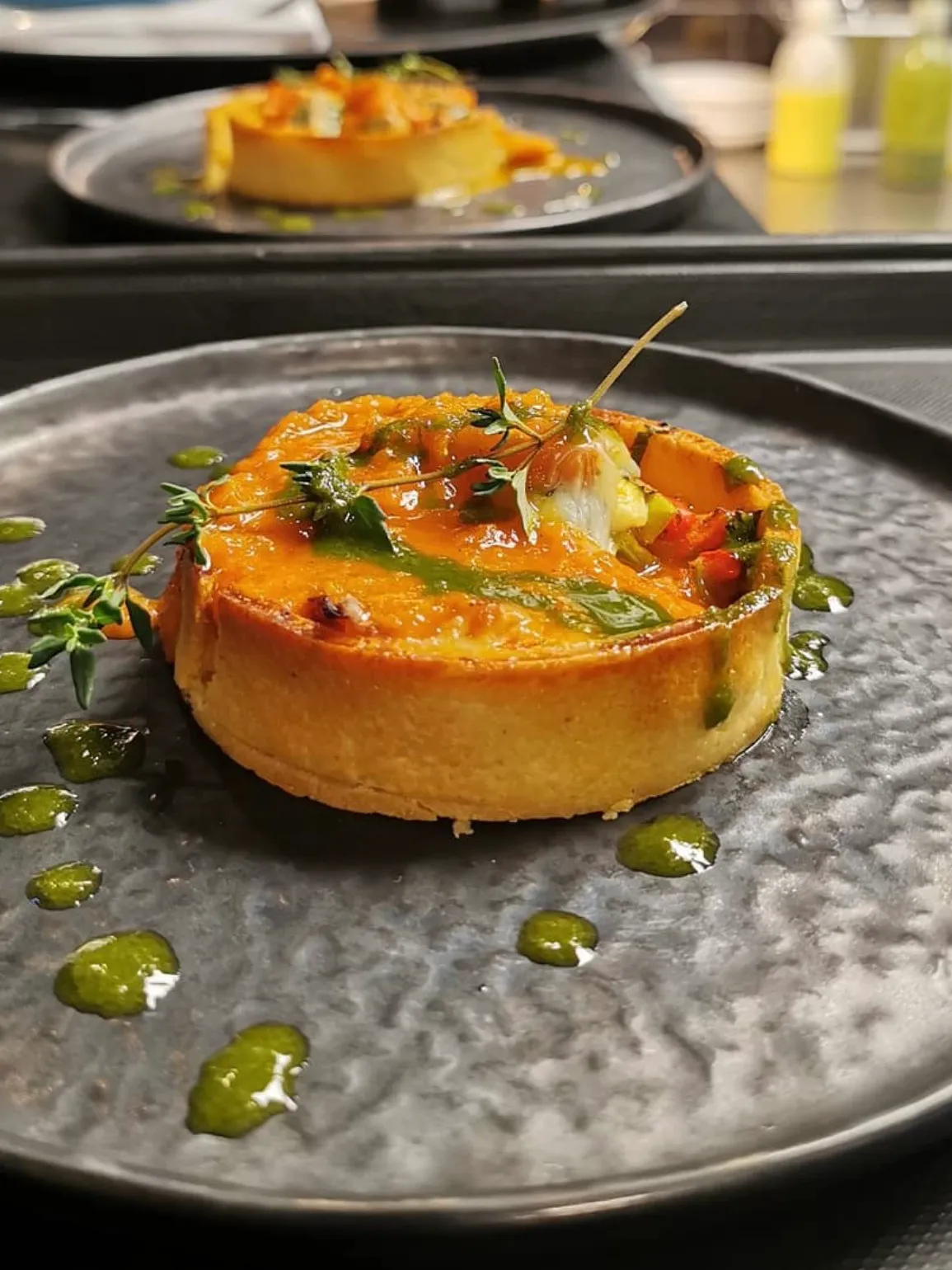 Lanthe Restaurant'ın zarif sunumuyla dikkat çeken, altın rengi kabuklu yuvarlak bir kiş veya tartın üzerinde turuncu soslu sebzeler, taze kekik dalları ve etrafına özenle serpiştirilmiş parlak yeşil yağ damlacıkları bulunan, şık koyu gri bir tabakta servis edilmiş bir yemek.