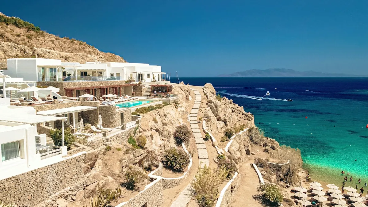 Mikonos'ta kayalık bir yamaca özenle inşa edilmiş, bembeyaz mimarisiyle dikkat çeken lüks bir tesis, berrak turkuaz renkteki Ege Denizi'ne nazır uzanıyor ve yüzme havuzları, şezlonglarla dolu terasları ve aşağıda hareketli bir kumsal sunarak unutulmaz bir tatil deneyimi vaat ediyor.