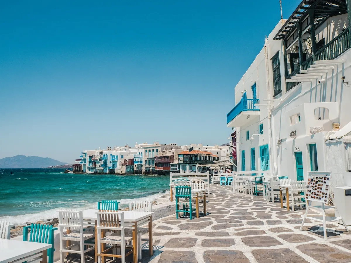 Mikonos adasının berrak turkuaz sularının kıyısında, mavi panjurlu ve balkonlu bembeyaz binaların sıralandığı pitoresk bir koyda, ön planda güneşin altında boş masaların ve sandalyelerin bulunduğu bir sahil restoranı alanı.