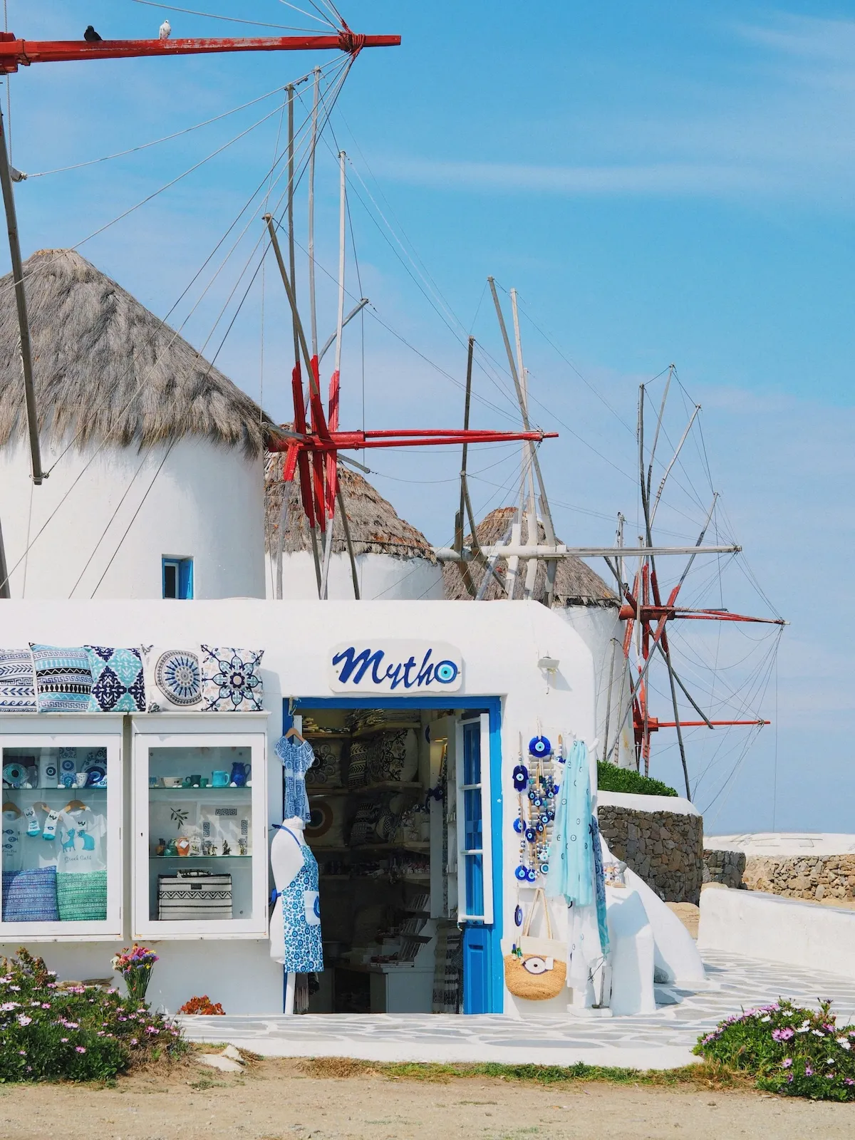 Mykonos'un simgesi olan, hasır çatılı ve kırmızı yelkenli geleneksel yel değirmenlerinin önünde konumlanan, Mytho yazılı ve mavi detaylı tabelası olan beyaz badanalı bir hediyelik eşya dükkanının vitrininde çeşitli seramikler ve nazarlıklar dikkat çekerken, berrak mavi gökyüzü bu turistik manzarayı aydınlatıyor.