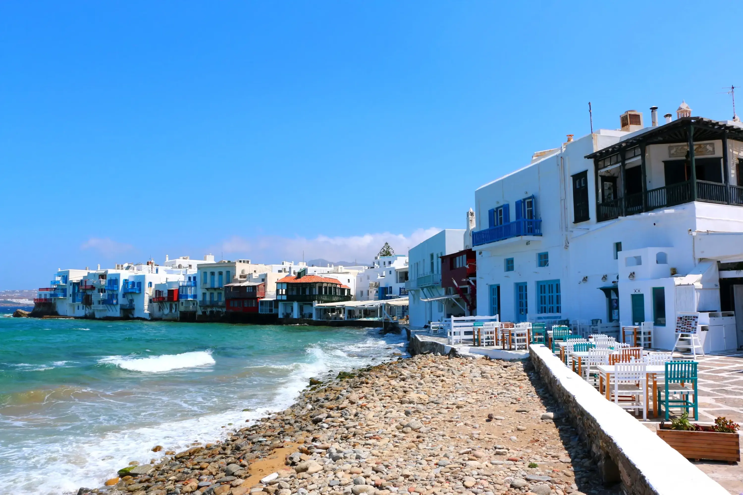 Mikonos'un canlı sahil kasabalarından birinde, turkuaz renkli denizin köpüklü dalgalarının çarptığı kayalık kıyı şeridi boyunca uzanan, karakteristik mavi kepenkli ve beyaz badanalı evlerin ve keyifli açık hava restoranlarının bulunduğu pitoresk bir manzara.