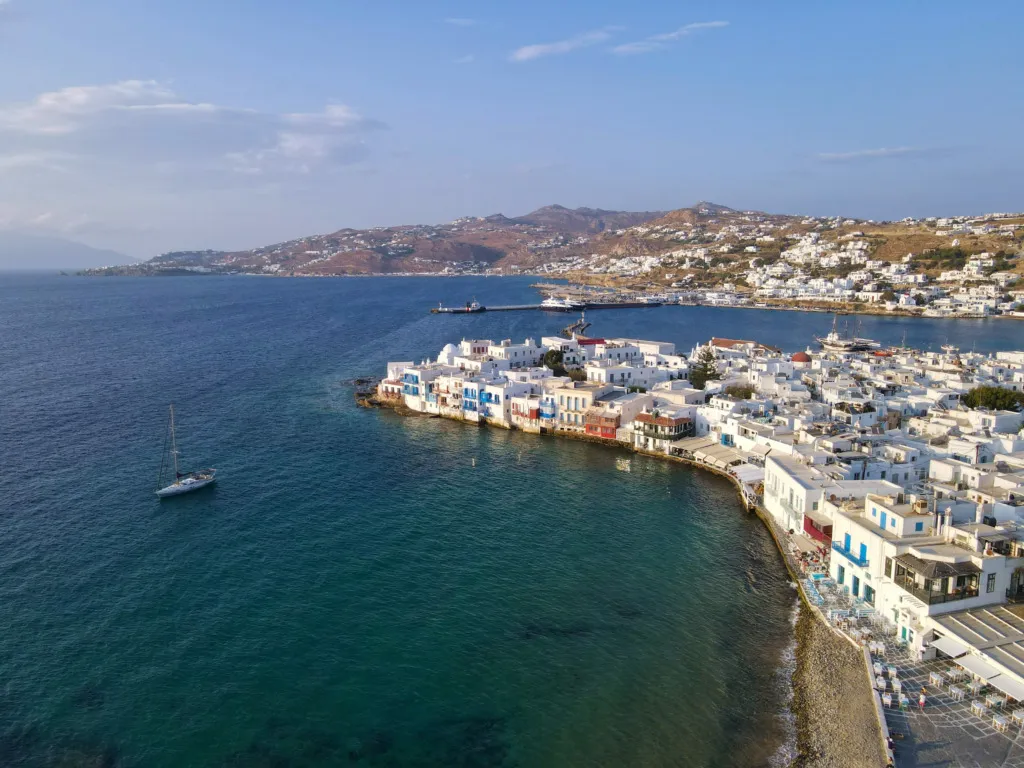 Mikonos'un en güzel plajları ve berrak turkuaz suları ile meşhur kıyı şeridinde yer alan bembeyaz evleri, denizde zarifçe ilerleyen bir yelkenli ve arka planda uzanan tepelerle çevrili eşsiz ada atmosferini yansıtan büyüleyici bir genel görünüm.
