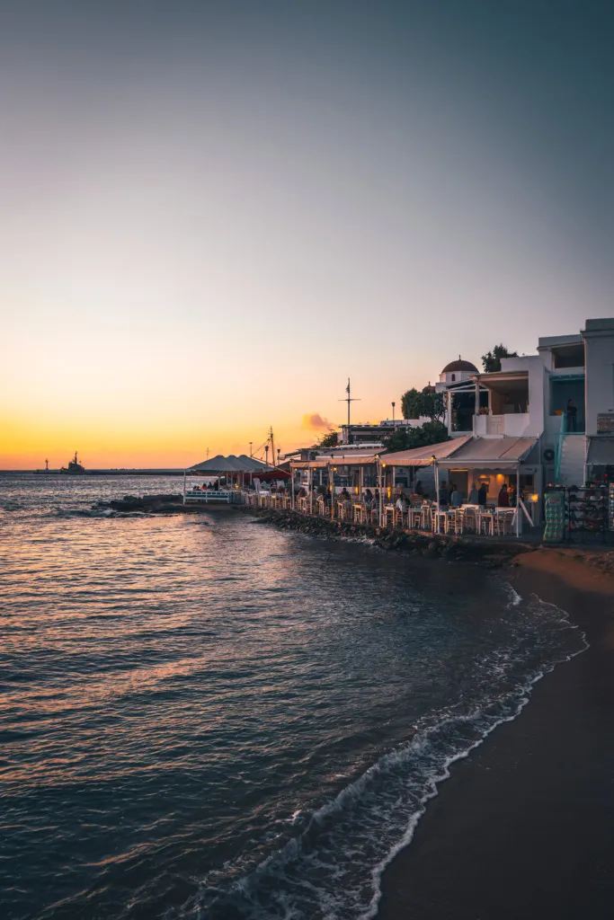 Mikonos sahilinde, batan güneşin turuncu ve mor renklerine bürünen gökyüzünün altında, denize sıfır uzanan beyaz cepheli tavernaların ve restoranların ışıkları yanarken, masalarda oturan misafirler eşsiz Akdeniz atmosferinin tadını çıkarıyor, hafif dalgaların kıyıya vurduğu kumlu plaj huzurlu bir akşamüstü manzarası sunuyor.