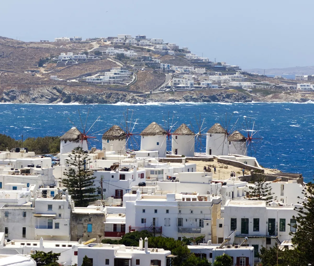 Mikonos adasının ünlü bembeyaz yel değirmenleri, saman çatılı ve kırmızı pervane detaylarıyla, dalgalı Ege Denizi'nin derin mavi sularına yukarıdan bakarken, altlarında yoğunlaşan beyaz badanalı geleneksel Kiklad evleriyle tepelik bir kıyı şeridini süslüyor.