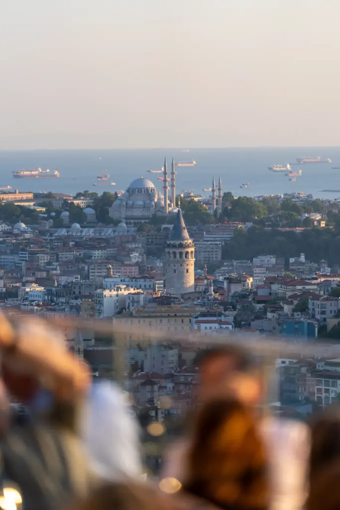Ön plandaki bulanık insan silüetlerinin ve teras camının ardından, tarihi Galata Kulesi, görkemli Süleymaniye Camii ve gemilerle süslenmiş uçsuz bucaksız Boğaz'ın eşsiz İstanbul manzarası, şehrin en iyi teraslı mekanlarından birinin sunduğu büyüleyici atmosferi yansıtıyor.