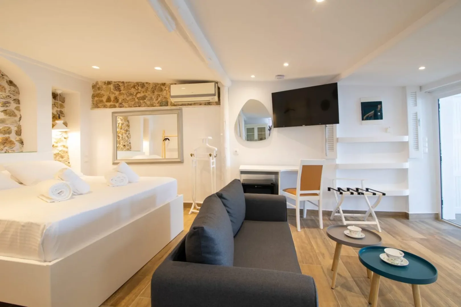 Pontikonisi'deki bir otel odasının modern tasarımı; beyaz çarşaflı bir yatak, rahat koyu gri bir kanepe, duvara monte edilmiş düz ekran TV, dekoratif aynalar, taş duvar detayları, beyaz raflar ve balkon kapısının yanında şık sehpalardaki çay setleriyle konforlu bir konaklama alanı sunuyor.
