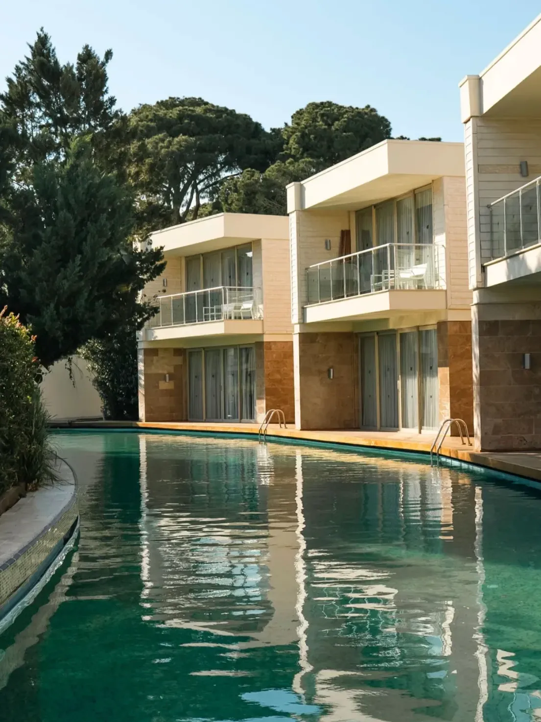Rixos Premium Belek'teki modern mimariye sahip lüks villaların veya otel odalarının, geniş ve berrak bir yüzme havuzuna bakan cam balkonları ve alt katlardaki doğrudan havuz erişimi, yeşil ağaçlarla çevrili doğal ve huzurlu bir tatil ortamı sunuyor.