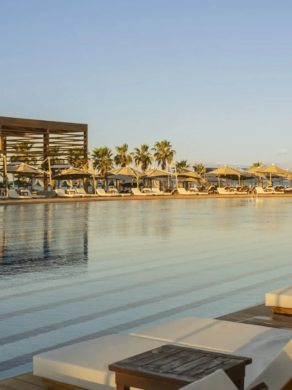 Şezlong ve şemsiyelerle çevrili, Belek'teki Rixos Premium otelinin berrak, çizgili yüzme havuzu, palmiye ağaçlarıyla süslenmiş geniş bir terası ve ahşap bir pergolayı yansıtarak zarif bir dinlenme ortamı sunuyor.