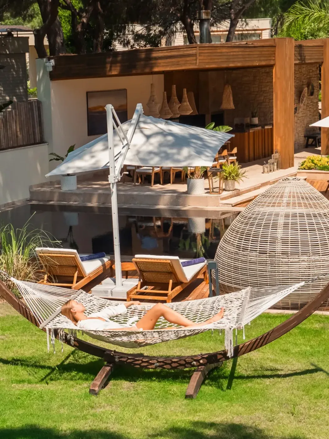 Rixos Premium Belek otelinin yemyeşil bahçesinde, beyaz bir hamakta uzanmış genç bir kadın, arkasında şık bir yüzme havuzu, ahşap teras, büyük bir şemsiye ile gölgelenmiş yemek alanı ve tropikal bitkilerle çevrili lüks bir ortamda huzur içinde dinleniyor.