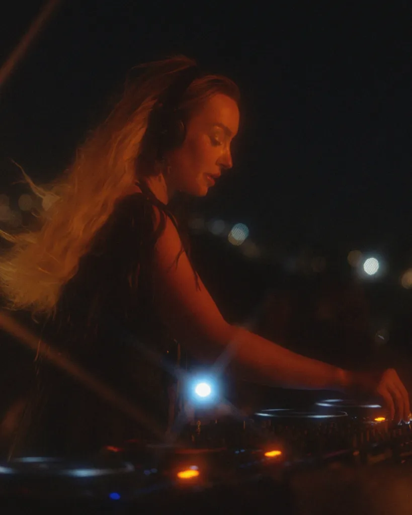 Sıcak turuncu bir ışıkla aydınlanan, kulaklıklarıyla DJ mikserinin başındaki bir kadın, gece karanlığında enerjik bir müzik performansı sergilerken, müziğin akışına kendini kaptırmış bir şekilde Scorpios'ta anı yakalıyor.