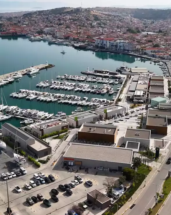 Bu etkileyici havadan görünüm, yüzlerce modern teknenin demirlediği geniş bir Setur marinasını, çevresindeki şık alışveriş ve dinlence tesislerini, otoparkları ve arka planda Ege kasabasının pitoresk konutlarını gözler önüne sererek canlı bir kıyı şeridi deneyimi sunuyor.