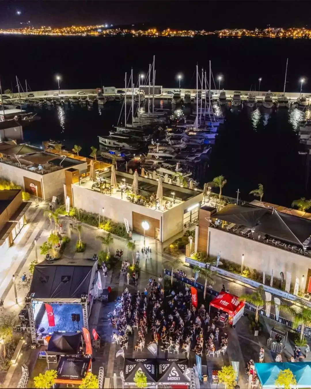 Işıl ışıl parlayan uzaktaki şehir ışıklarıyla çevrili bir koyda, çok sayıda yat ve teknenin demirli olduğu bir marina, hemen yanında ise modern binalar, palmiye ağaçları ve açık hava sahnesinde düzenlenen etkinliği izleyen geniş bir kalabalığın bulunduğu hareketli bir gece manzarası.