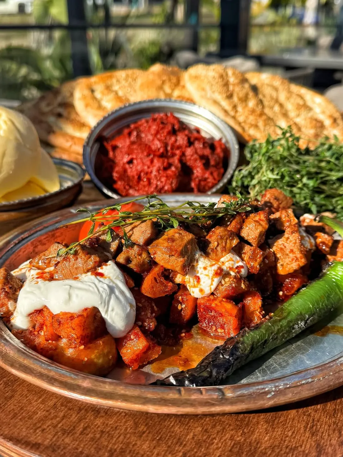 Sıralı kebap servis edilen geniş bakır tepside, üzerine yoğurt gezdirilmiş, kızarmış et küpleri ve sebzelerden oluşan ana yemeğin yanında közlenmiş yeşil biber, arka planda ise kırmızı biber salçası, pide ekmeği ve tereyağı yer alıyor, zengin bir Türk ziyafeti sunuluyor.