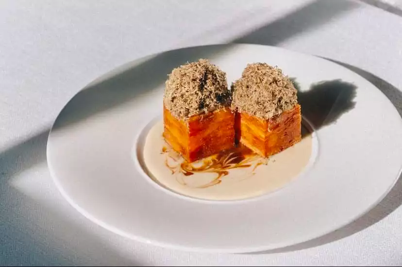 Urla Vino Locale restoranının menüsünden, güneşin gölgeler oluşturduğu beyaz bir masada zarifçe sunulan, kremsi bir sosun üzerinde yer alan ve bolca rendelenmiş trüf mantarı ile taçlandırılmış iki adet katmanlı, altın rengi, gurme lezzetin yakın çekim fotoğrafı.