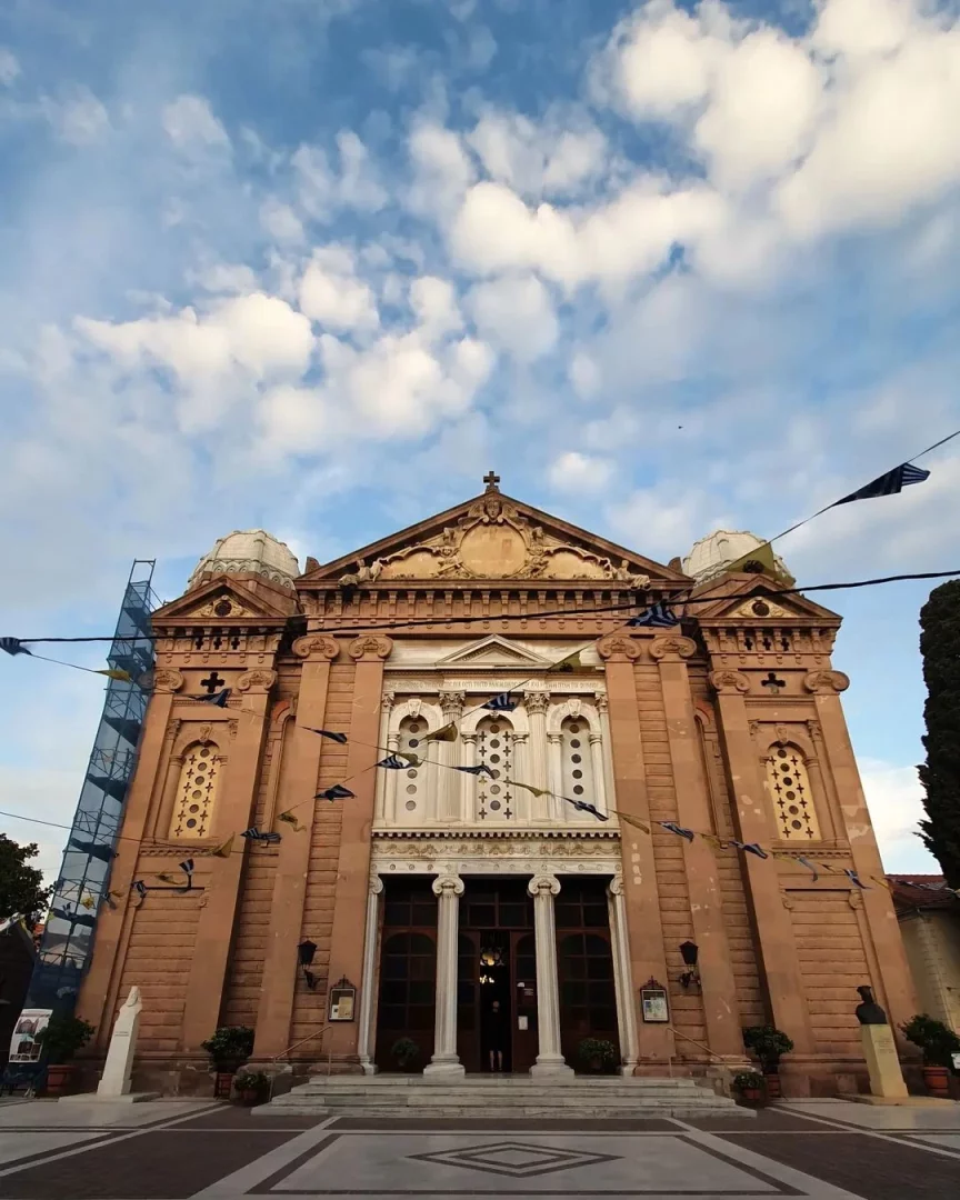Agios Therapon Kilisesi'nin görkemli kırmızımsı kahverengi cephesi, sütunlu ana girişi, haçla taçlanmış alınlığı ve dekoratif pencereleriyle, sol tarafında restorasyon iskelesi ve önünde mavi-beyaz bayrak süslemeleriyle, altında geometrik desenli geniş bir meydanla bulutlu gökyüzünün altında yükseliyor.