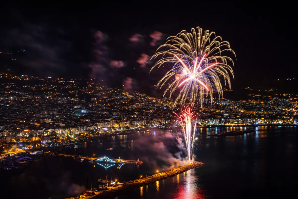 Alanya'nın hareketli ve ışıl ışıl gece şehir manzarası üzerinde gökyüzünü aydınlatan rengarenk havai fişekler, Akdeniz'in sakin sularına yansıyarak büyüleyici bir görsel şölen sunarken, sahil şeridi boyunca uzanan binalar da aydınlık bir fon oluşturuyor.