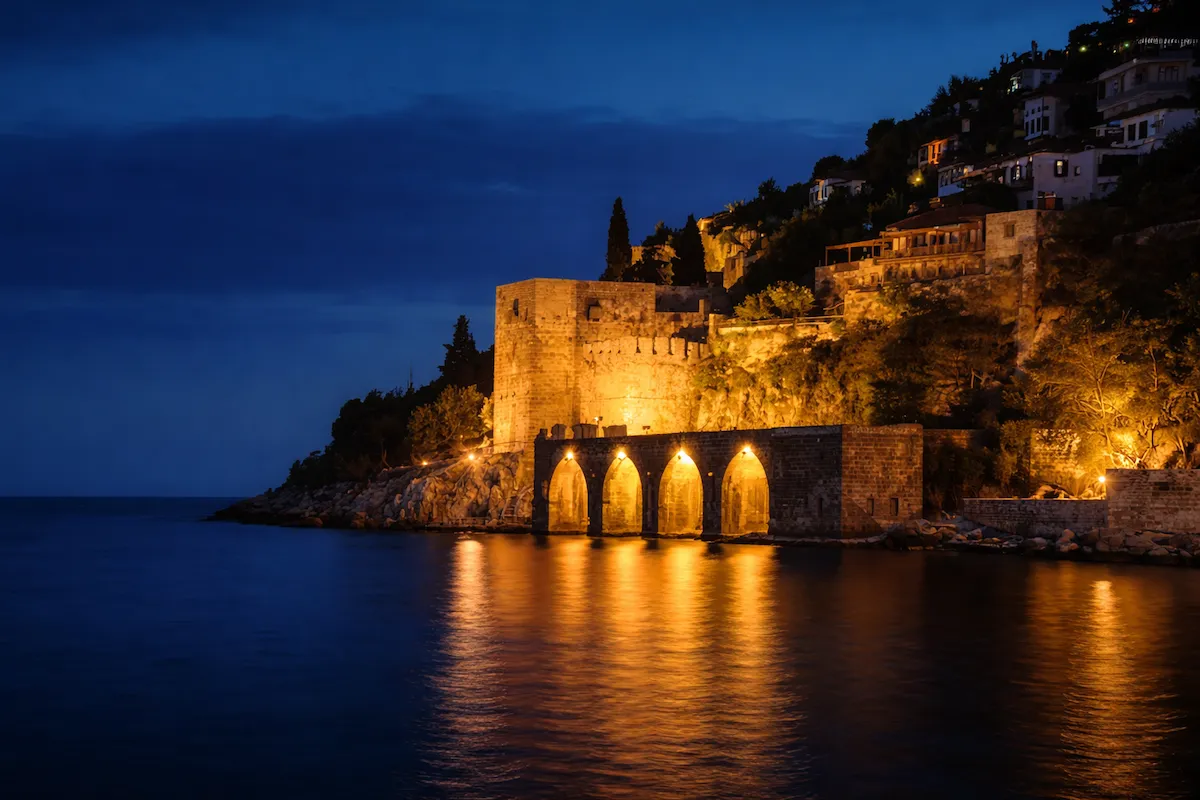 Alanya Kalesi sur duvarlarının ve tarihi Alanya Tersanesi'nin deniz kenarında, gece karanlığında parlayan sıcak ışıklarla aydınlatıldığı, suyun üzerinde büyüleyici yansımalar oluşturan pitoresk bir manzara.