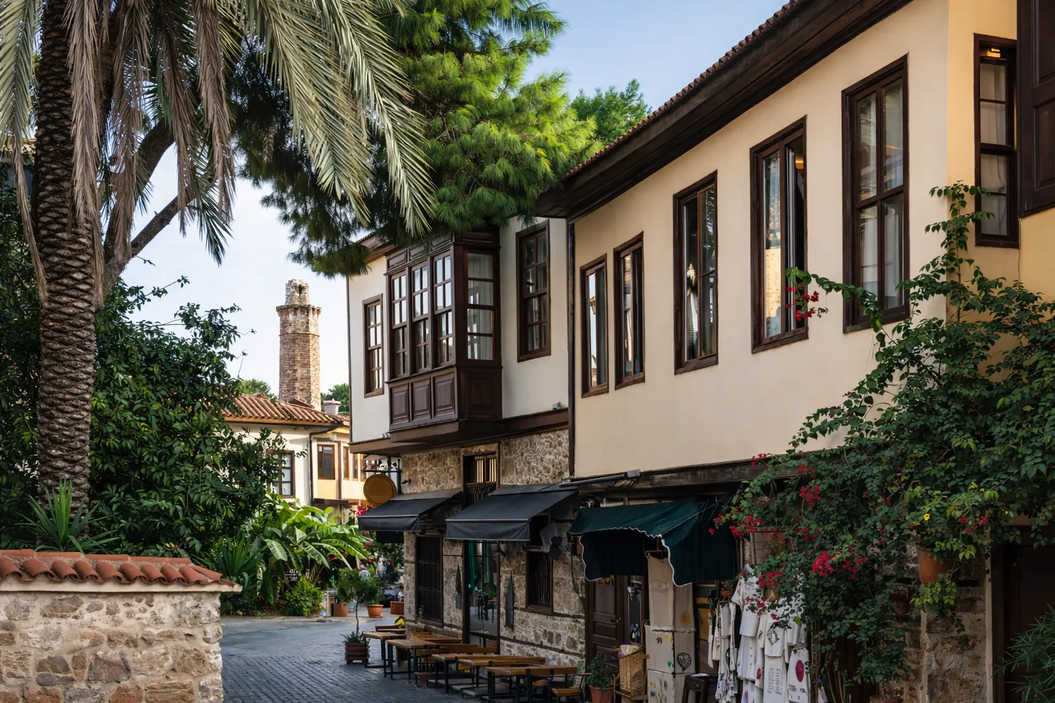 Antalya'nın tarihi Kaleiçi'nde, yemyeşil ağaçlar ve palmiyelerle çevrili geleneksel evlerin ve küçük dükkanların sıralandığı Arnavut kaldırımlı bir sokak boyunca, uzakta meşhur Kesik Minare Camii'nin taş minaresi görünüyor.