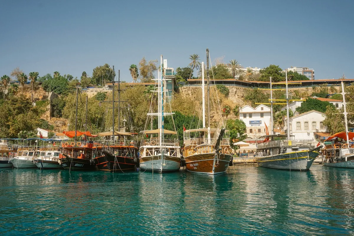 Antalya'nın hareketli yat limanında, turkuaz renkli berrak denizin üzerinde sıralanmış, geleneksel tasarımlı çok sayıda gezinti teknesi ve arka planda yeşil bitki örtüsüyle kaplı dik falezler ile tarihi yapılar güneşli bir günde büyüleyici bir kıyı manzarası oluşturuyor.