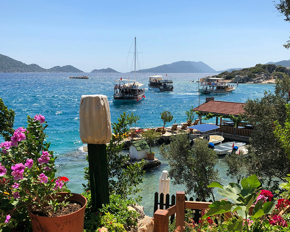 Güneşli bir Akdeniz gününde, yemyeşil tepelerle çevrili berrak turkuaz sularında seyreden tur tekneleri ve kıyıda begonvil çiçekleriyle süslenmiş bir restoranın görüldüğü bu manzara, Antalya seyahat rehberinin denizden keşfedilecek koylarına dair davetkar bir izlenim sunuyor.