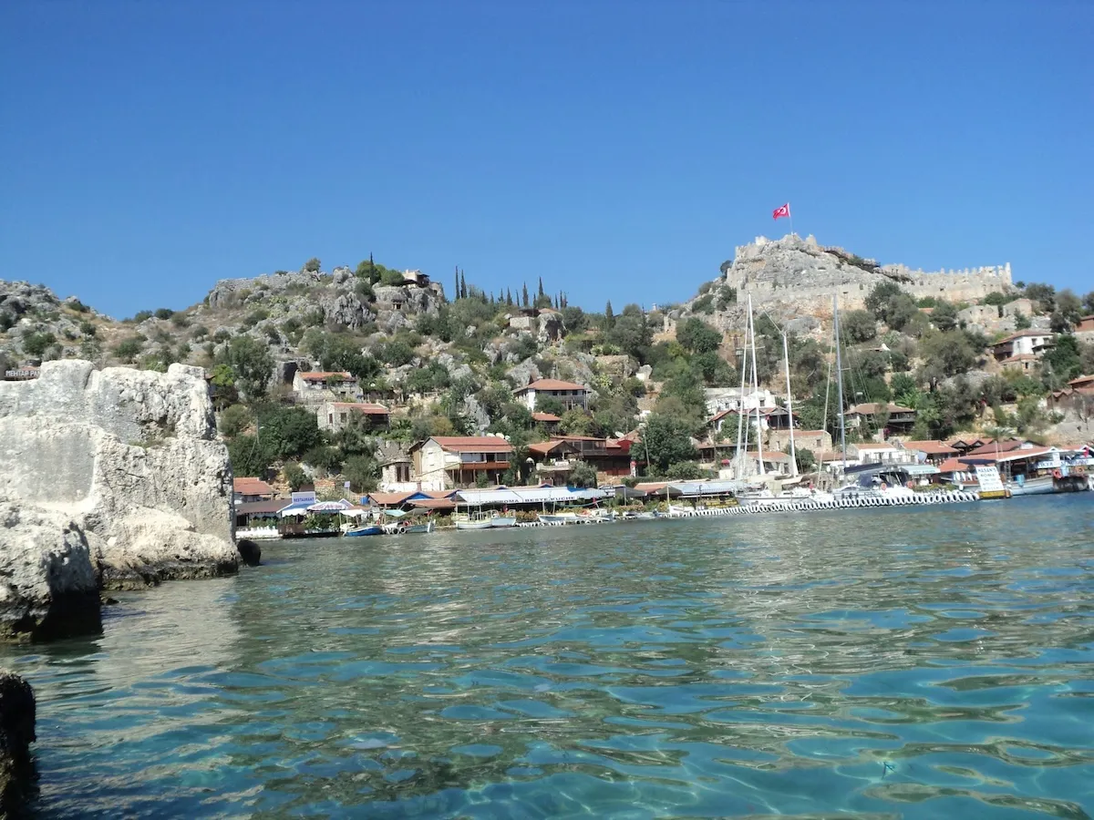 Antalya'nın berrak turkuaz sularına nazır, kayalık yamaçlara yayılmış kırmızı çatılı evleri, teknelerle dolu hareketli limanı ve zirvesinde Türk bayrağı dalgalanan sarp bir antik kaleyi içeren bu pitoresk koy, tipik bir Akdeniz sahil kasabasının ruhunu yansıtıyor.