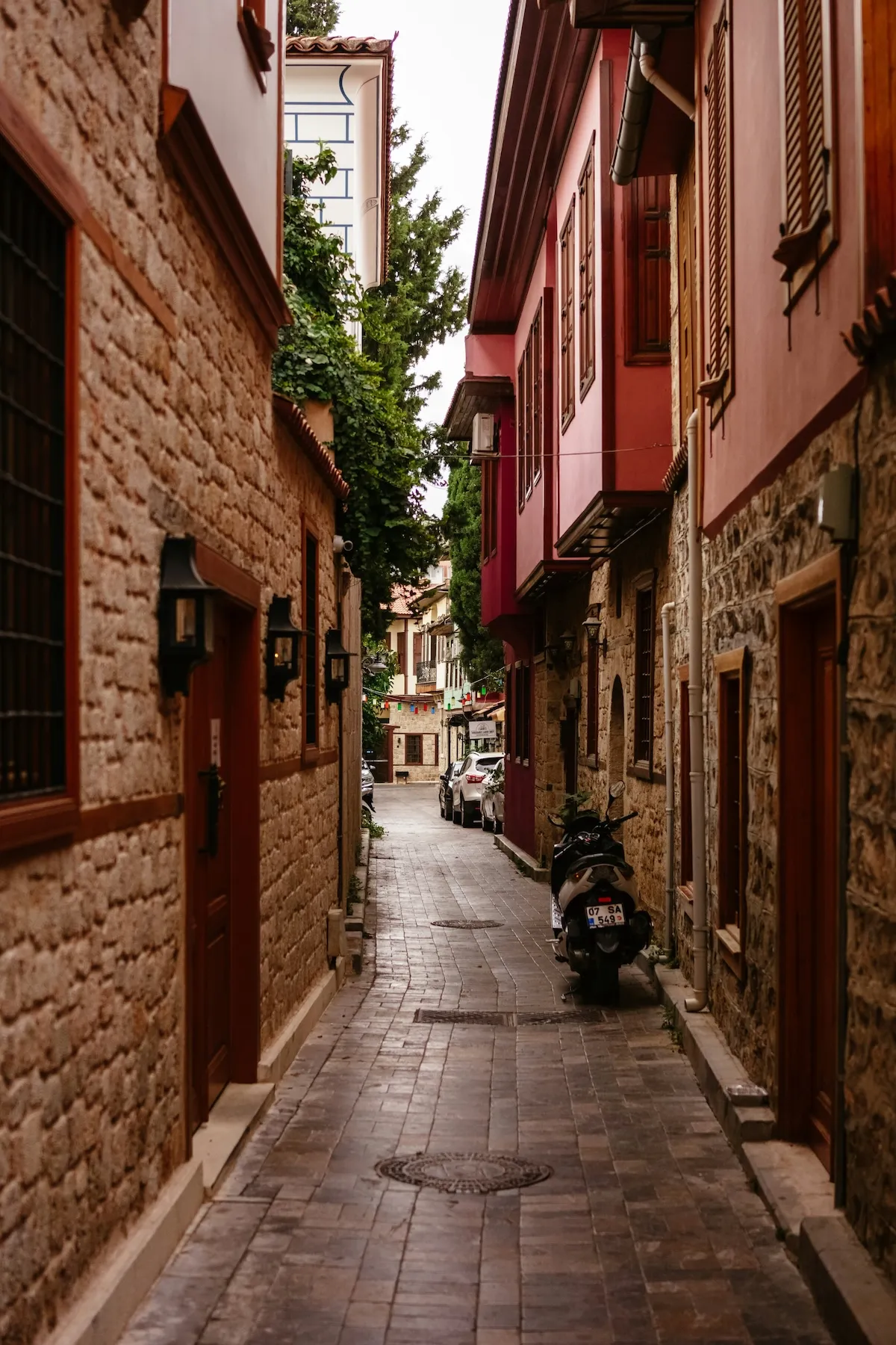 Antalya'nın dar sokaklarından birinde, tarihi dokusunu yansıtan taş duvarlı ve ahşap pencereli, bazıları kırmızımsı tonlarda boyalı geleneksel evlerin sıralandığı Arnavut kaldırımlı bir yolda, sağda park etmiş bir motosiklet ve sokağın ilerisinde sıralanmış birkaç araba görülüyor.
