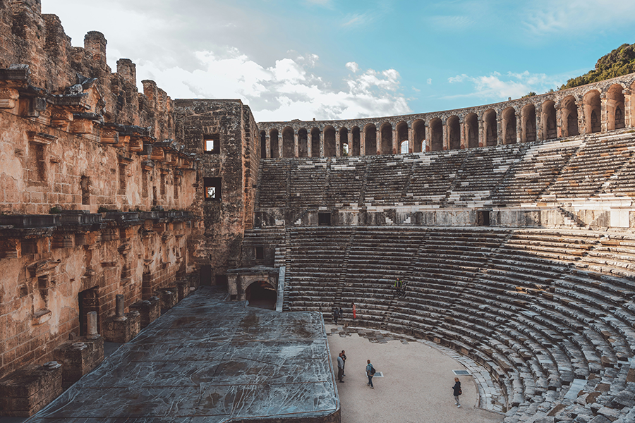 Antalya gezi: Antalya'da bulunan Aspendos Antik Tiyatrosu'nun iyi korunmuş taş yapısı, çok katlı sahne binası ve geniş oturma sıralarını saran kemerli galerisi ile mavi gökyüzü altında duran görkemli hali, sahne önünde dolaşan birkaç ziyaretçiyle birlikte kuşbakışı bir açıdan çekilmiş.