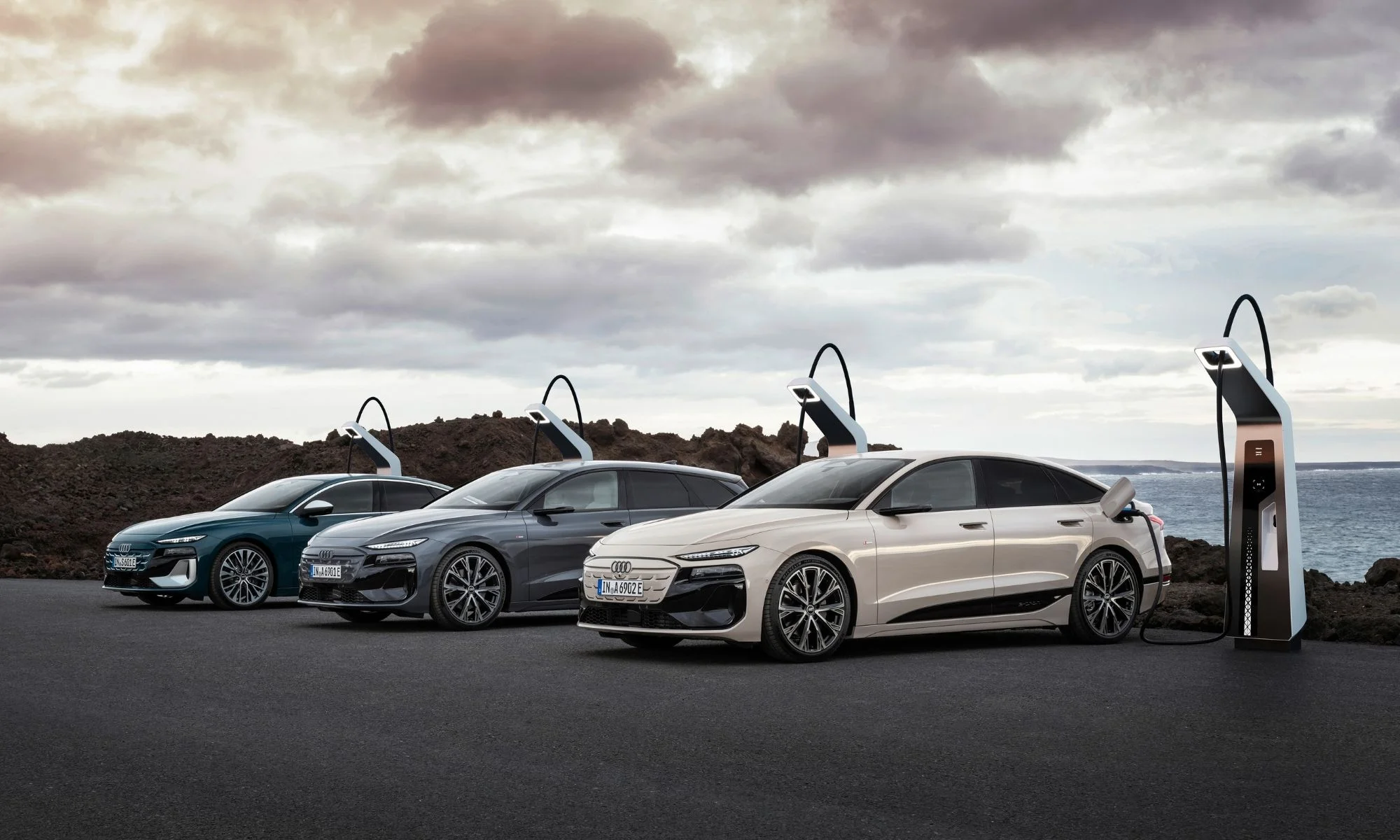Bulutlu gökyüzü ve dalgalı denizin görüldüğü kayalık bir arazinin kenarında, açık krem rengi bir Audi A6 e-tron ile iki gri ve yeşil Audi A6 e-tron Avant modeli, modern şarj istasyonlarında düzenli bir sıra halinde şarj edilmektedir.