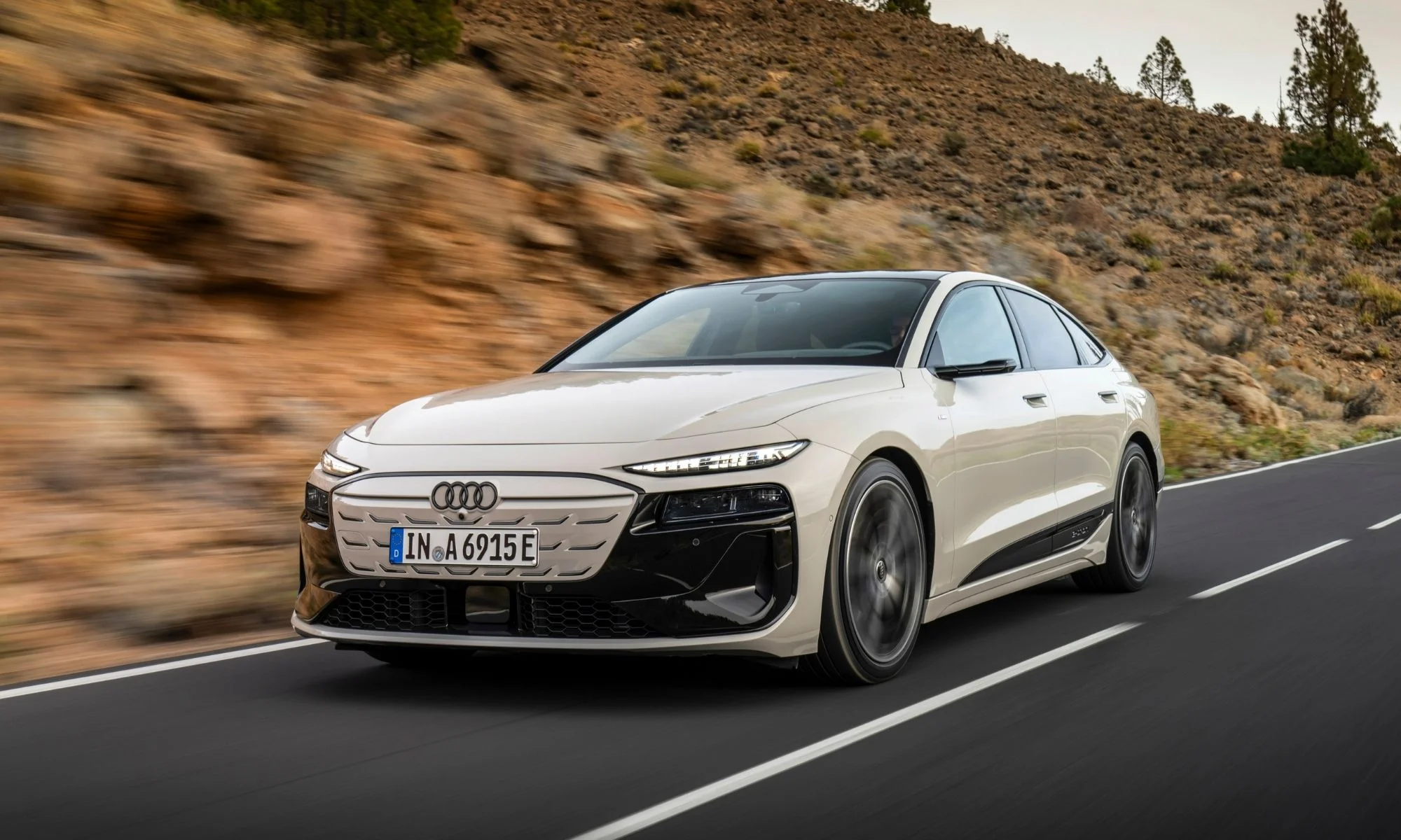 Bej kum rengi ve siyah detaylara sahip şık Audi A6 e-tron konsept otomobil, hızla ilerlediği belli olan yolda, arka plandaki kahverengi kayalıklar ve bulanık ağaçlar eşliğinde dinamik ve modern bir görünüm sunuyor.