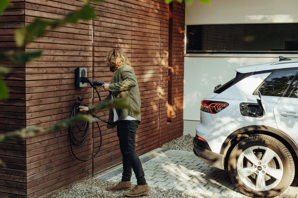 Modern ahşap cepheli bir evin önündeki gri taşlı zeminde duran beyaz Audi e-tron elektrikli otomobili, uzun saçlı bir adam duvardaki ev tipi şarj istasyonuna takılı kabloyu aracın şarj portuna özenle bağlayarak şarj ediyor.