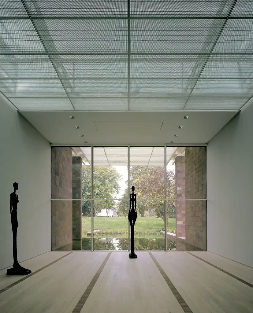 Fondation Beyeler'in geniş ve minimalist bir galeri odasının iç görünümü. Odada iki uzun, ince, koyu renkli heykelsi figür bulunuyor; biri solda, diğeri merkezde. Arka planda büyük cam pencereler, sakin bir su havuzu ve yemyeşil ağaçlarla çevrili bir park manzarasını gösteriyor. Tavan metalik bir ızgara yapısına sahip.