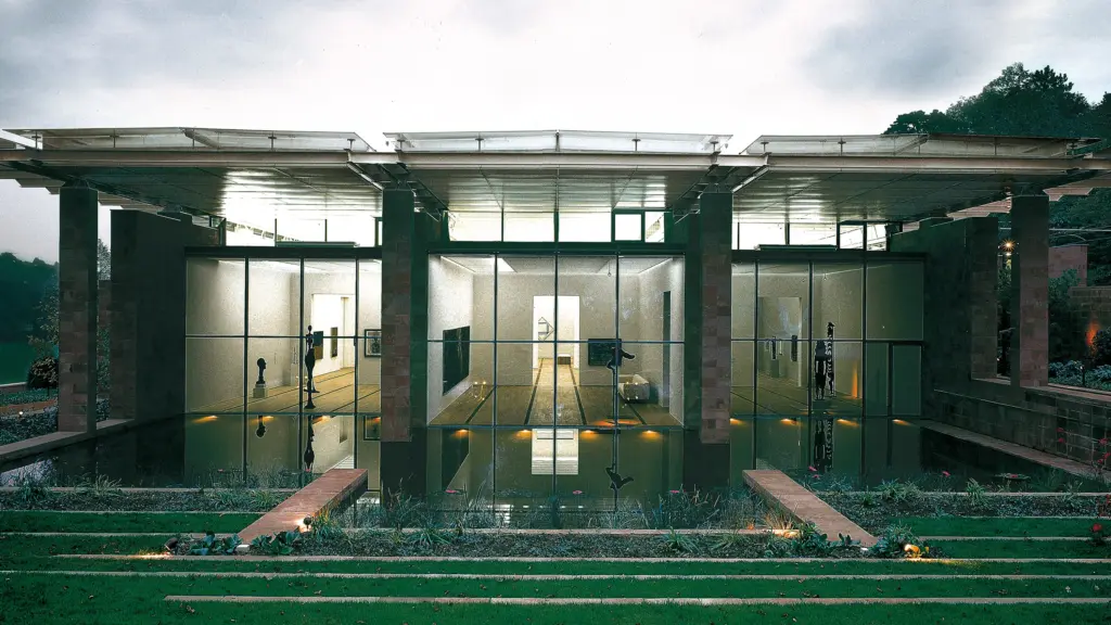 Fondation Beyeler müzesinin dış cephesinin geniş bir görünümü, yatay çizgili ve cam panellerden oluşan modern mimarisiyle dikkat çekiyor. Binanın önünde, heykellerin yansımalarının görülebildiği bir yansıtma havuzu var. Havuzun çevresinde terracota renkli duvarlar ve ön planda basamaklı yeşil çim teraslar bulunuyor.