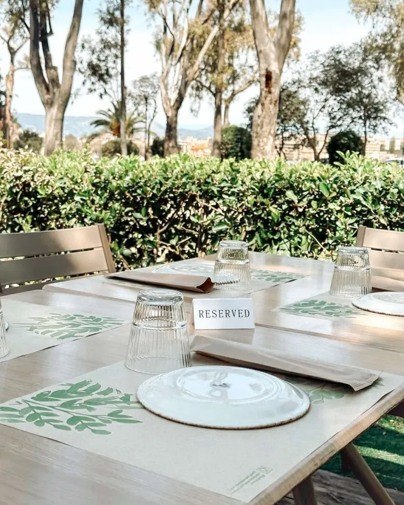 Avli Korfu restoranının yemyeşil ağaçlar ve çitlerle çevrili, dağ manzaralı açık hava bölümünde, üzerinde Rezerve kartı bulunan, yaprak desenli Amerikan servisleri, ters duran bardaklar ve beyaz tabaklarla özenle hazırlanmış ahşap bir masa gözüküyor.