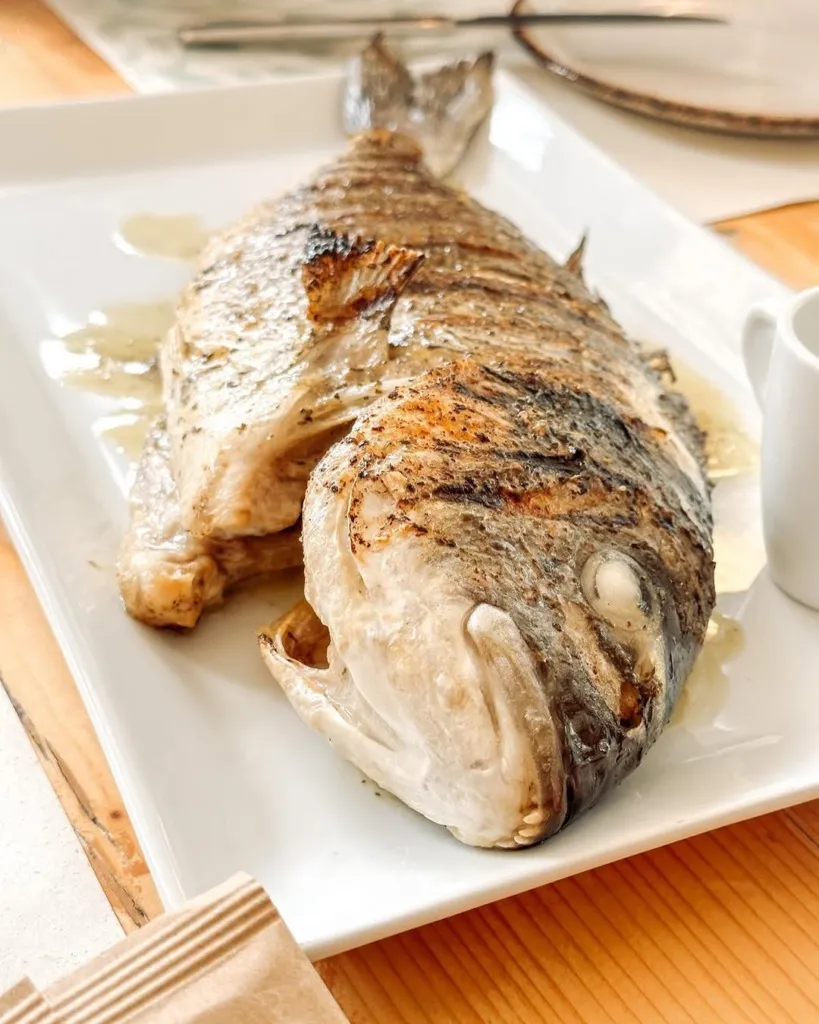 Avli Korfu Restoranı'nda servis edilen, hafifçe zeytinyağı gezdirilmiş gibi duran ve belirgin ızgara izleriyle dikkat çeken, başı ve kuyruğuyla bütün olarak sunulmuş taze balık, beyaz dikdörtgen bir servis tabağında ahşap bir masanın üzerinde, yanında küçük bir sos sürahisiyle iştah açıcı bir görünüm sergiliyor.