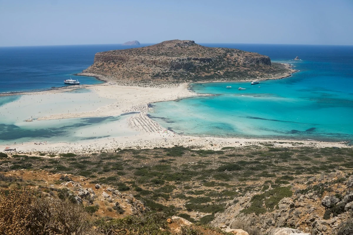 Girit'teki Balos Plajı'nın tepeden geniş açılı görünümünde, beyaz kumlu bir kumsal, sığ turkuaz sularla çevrili kayalık bir yarımadayı anakaraya bağlıyor; plajda ve lagün sularında çok sayıda tatilci, kumsal boyunca sıralanmış şemsiyeler ve uzakta demirlemiş tekneler net bir şekilde izleniyor.
