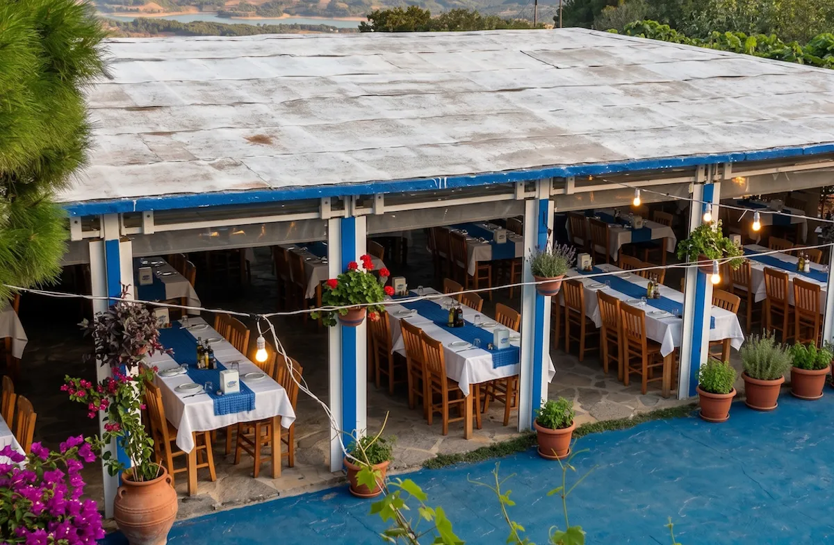 Tepeköy'deki Barba Yorgo Taverna'nın mavi-beyaz direkleri, zarif asma lambaları ve uzaklardaki göl manzarasıyla çevrili, saksı çiçekleriyle süslü, bembeyaz masa örtüleri ve lacivert runner'larla donatılmış masaların bulunduğu geniş ve davetkar açık hava yemek alanı.