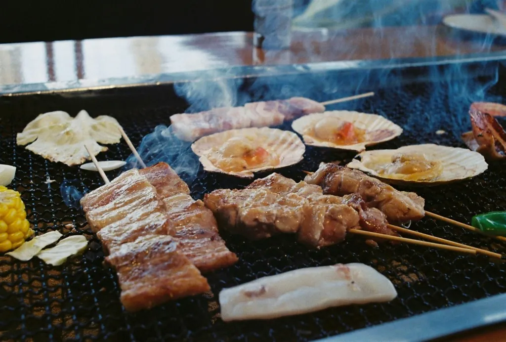 Mısır, mantar, et şiş ve deniz tarağı gibi çeşitli yiyeceklerin mangalda közlenerek pişirildiği dumanlı bir barbekü sahnesi.
