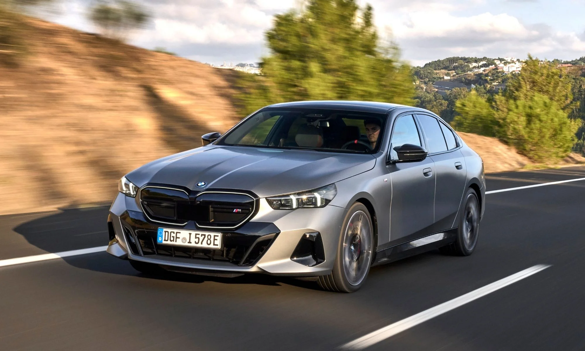 Gri bir BMW i5 elektrikli 5 Serisi, direksiyonunda bir sürücüyle birlikte, belirgin M Sport ön ızgarası ve parlak farlarıyla virajlı bir yolda dinamik bir şekilde ilerlerken, arkasında yeşil ağaçlar ve kuru tepeler görülüyor.
