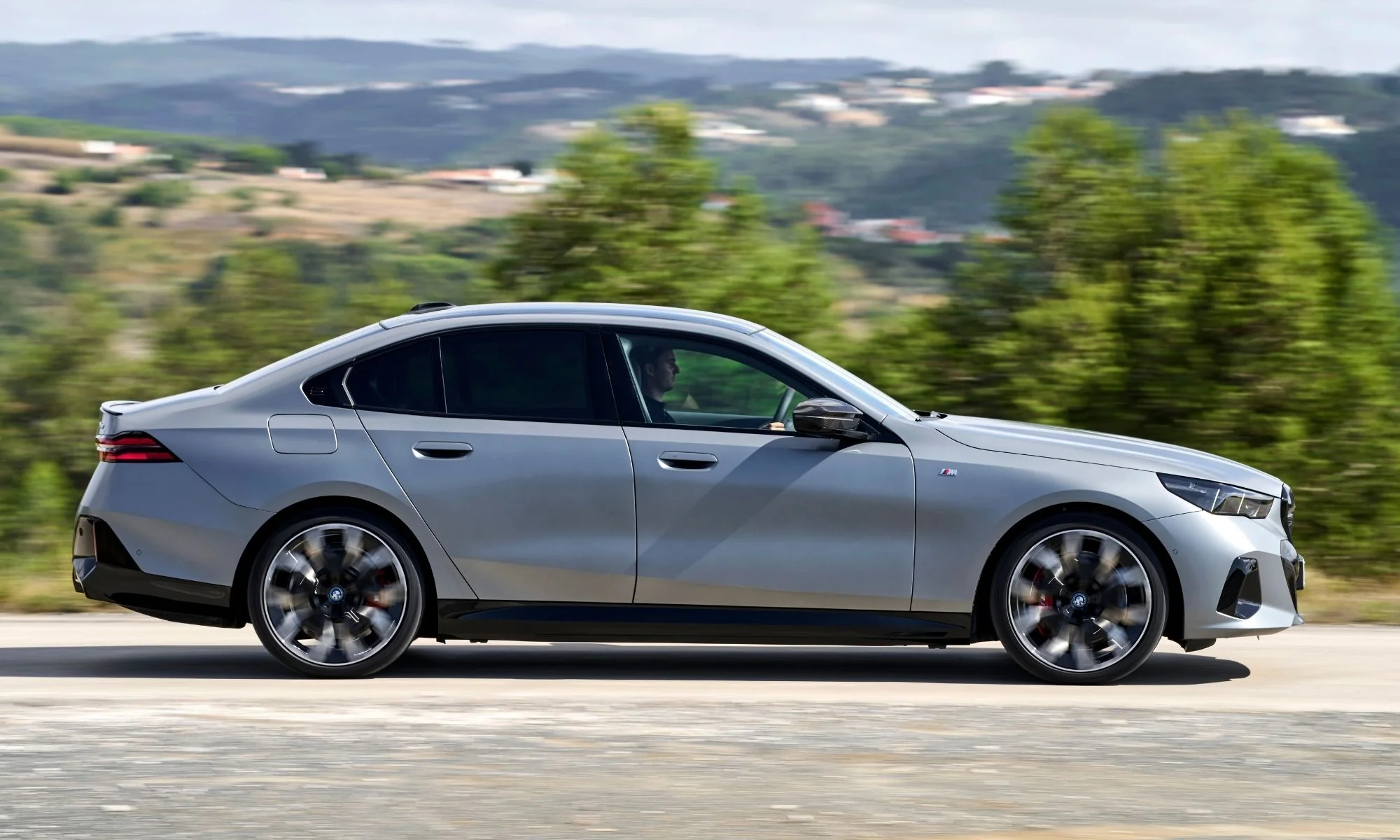 Gümüş gri renkteki yeni BMW i5 Elektrikli 5 Serisi, yeşil ağaçlarla kaplı tepelerin önünden hızla geçerken yan profilden görülen dinamik tasarımı ve akıcı hatlarıyla dikkat çekiyor.
