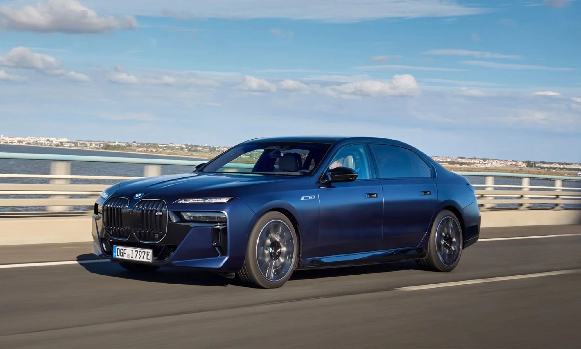 Parlak lacivert rengindeki yeni nesil BMW i7 M Sport Shadow Paket, geniş ve karanlık ön ızgarasıyla bir köprü üzerinde ilerlerken, arka planda mavilikler üzerinde uzanan şehir silueti ve hafif bulutlu gökyüzü dikkat çekiyor.