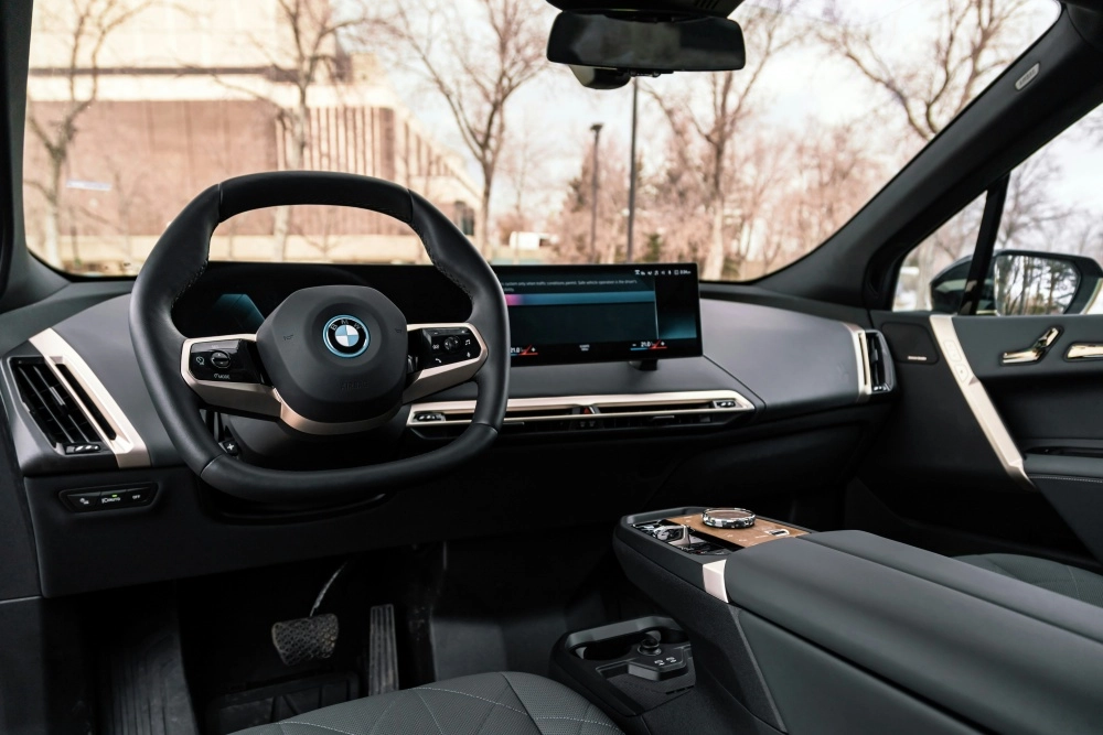 BMW iX elektrikli otomobilin modern iç mekanı, siyah direksiyon simidi, teknolojik geniş kavisli dijital ekranı, bronz detaylarla süslenmiş ön paneli ve şık orta konsolu ile sürücü koltuğundan panoramik bir görünüm sunuyor, dışarıda ağaçlar ve bir bina seçiliyor.